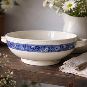 May include: A cream-coloured serving bowl with a blue floral band. The bowl has two handles and sits on a pedestal base. A vintage spoon rests on a folded linen napkin. Daisies and a white jug are in the background.