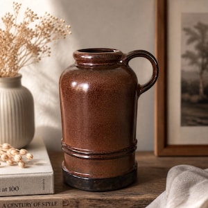 Può includere: Una brocca in ceramica marrone con manico. La brocca ha una superficie strutturata e una base marrone scuro. È su una superficie di legno. A sinistra c'è un piccolo vaso con fiori secchi.