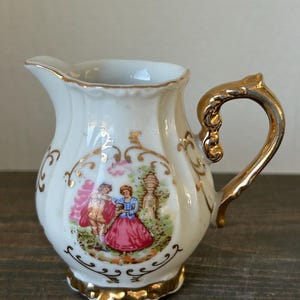 May include: A small, white porcelain creamer with a gold handle and trim. The creamer features a painted scene of a couple in period clothing. The handle and base are adorned with gold accents, adding a touch of elegance to the piece.