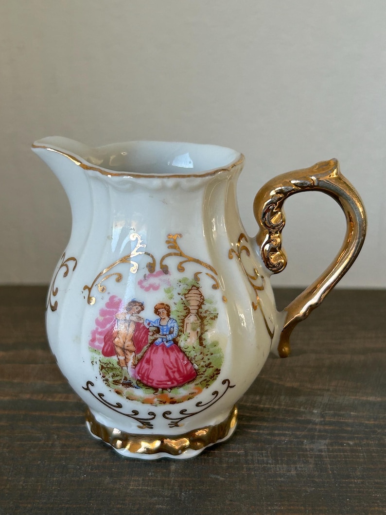 May include: A small, white porcelain creamer with a gold handle and trim. The creamer features a painted scene of a couple in period clothing. The handle and base are adorned with gold accents, adding a touch of elegance to the piece.