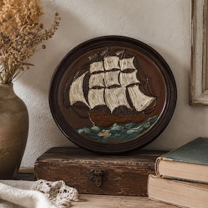 May include: A circular, brown wall plaque depicting a sailing ship in relief, with white sails and a brown hull. The ship sails on blue and green waves. The plaque is displayed on a wooden box.