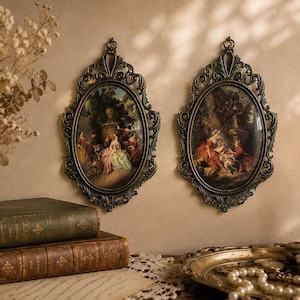 May include: Two oval wall frames with ornate bronze-coloured metal borders, each containing a detailed painting of figures in a garden setting. The frames feature intricate scrollwork and a hanging loop at the top, displaying a dark, aged finish.