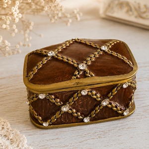 Pode incluir: Uma caixa de joias retangular castanha com um padrão de treliça em tom dourado. A superfície é decorada com pequenos strass redondos e transparentes incrustados em cada interseção da treliça de metal dourado, criando uma aparência texturizada e ornamentada.