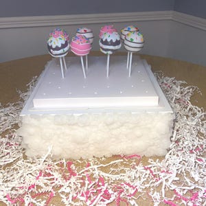 May include: A display of colorful cake pops on a white tiered stand. The cake pops have various toppings, including sprinkles and frosting. The stand is decorated with white fluffy material and surrounded by white and pink shredded paper.