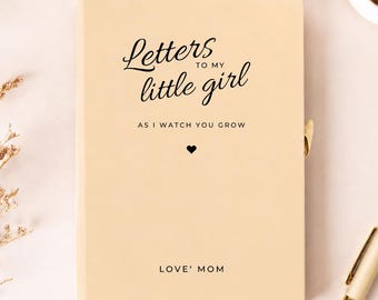 Può includere: Un diario color beige chiaro con testo nero che recita Letters TO MY little girl AS I WATCH YOU GROW con una piccola icona a forma di cuore e il testo LOVE' MOM in basso. Il diario ha una copertina liscia e una penna in metallo dorato è vicina.
