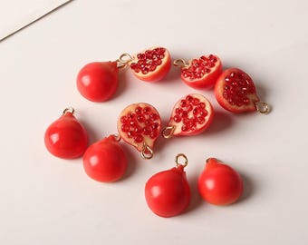 Può includere: Una collezione di ciondoli a forma di melograno rossi e ciondoli a metà melograno con anelli color oro. I ciondoli sono disposti su una superficie bianca. L'immagine include il testo "CG STUDIO CRAFT&GIFT DESIGN".