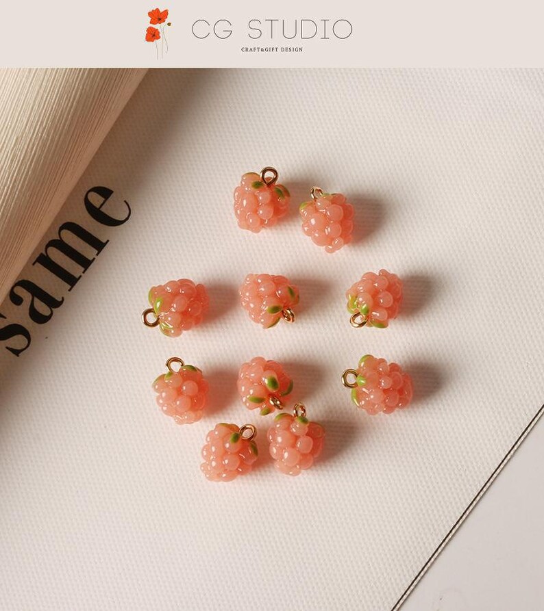 Pu&ograve; includere: Primo piano di diversi ciondoli a forma di lampone rosa con dettagli verdi e anelli dorati. I ciondoli sono sparsi su una superficie bianca. &Egrave; incluso anche il testo "CG STUDIO CRAFT&GIFT DESIGN".