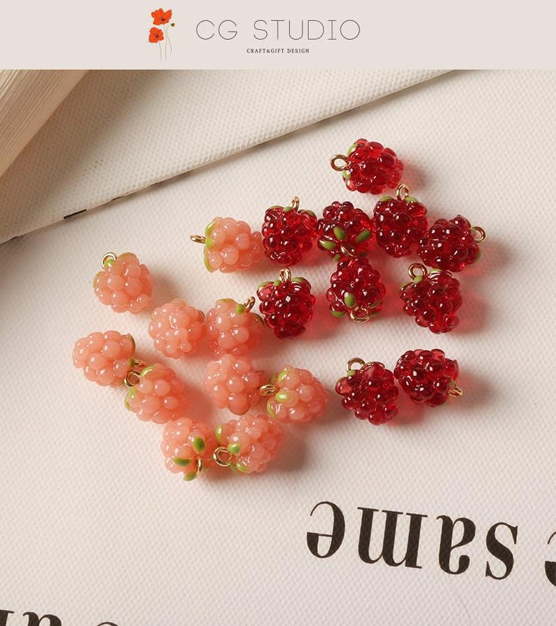 Pu&ograve; includere: Primo piano di diversi ciondoli a forma di lampone in varie tonalit&agrave; di rosso e rosa. I ciondoli hanno una finitura lucida e piccoli anelli color oro per il fissaggio ai gioielli. &Egrave; visibile anche il testo "CG STUDIO CRAFT&GIFT DESIGN".