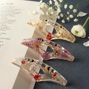 May include: Three clear resin book page holders with dried flowers and gold flakes. Each has a name: "Benjamin", "Chantal", and "Amelia". The holders also feature a moon phase design.