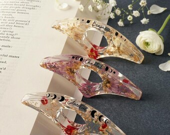 Può includere: Tre fermapagina in resina trasparente con fiori secchi e scaglie d'oro. Ognuno ha un nome: "Benjamin", "Chantal" e "Amelia". I fermapagina presentano anche un design a fasi lunari.