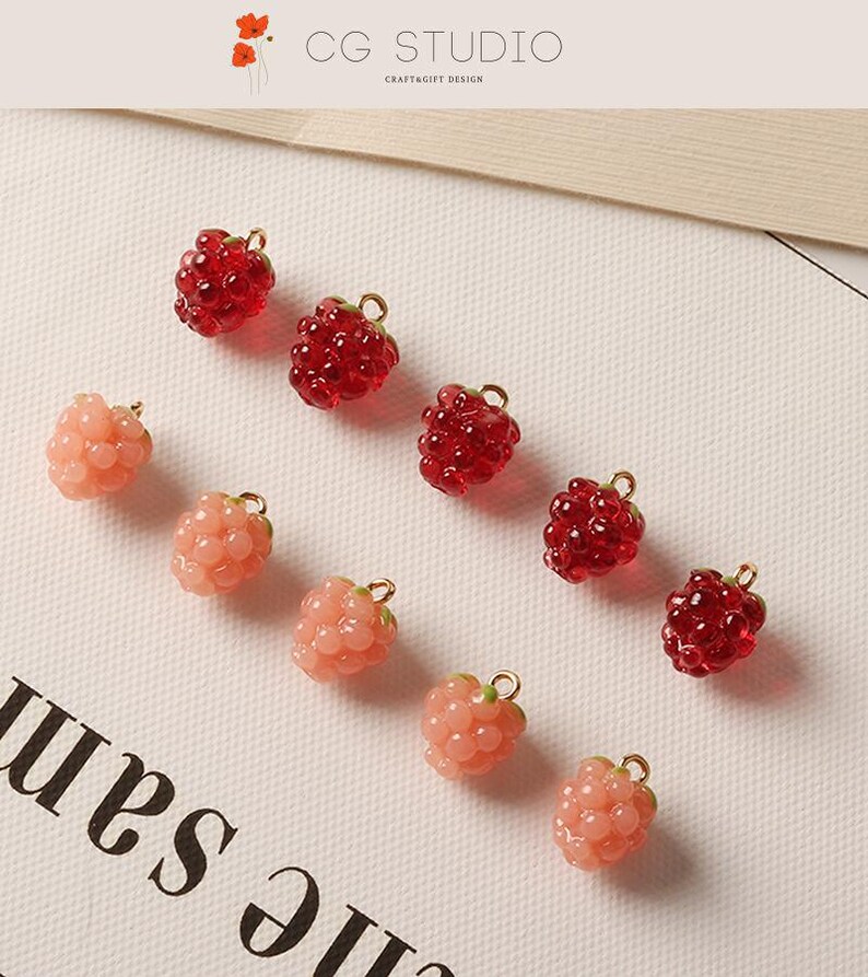 Pu&ograve; includere: Primo piano di diversi ciondoli a forma di lampone in varie tonalit&agrave; di rosso e rosa. Ogni ciondolo ha un anello color oro nella parte superiore. I ciondoli sono disposti su una superficie bianca. Il testo "CG STUDIO CRAFT&GIFT DESIGN" &egrave; visibile.
