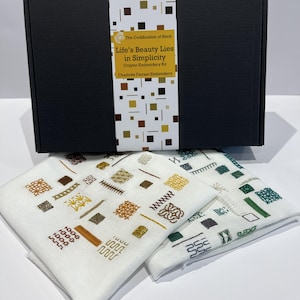 Peut inclure: Une boîte noire avec une bande décorative portant le texte "Life's Beauty Lies in Simplicity" et deux linges brodés blancs avec des motifs géométriques bruns, dorés et verts. Le kit est pour la broderie cryptique.