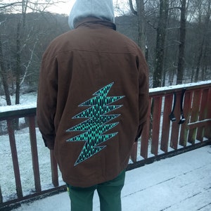 May include: A brown jacket with a green and black lightning bolt design on the back. The jacket is worn over a grey hooded sweatshirt. The person is standing on a snowy wooden deck.