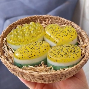 May include: A woven basket filled with handmade soaps. The soaps are various shapes, including a heart and oval, with yellow tops speckled with black dots. The soaps have a white and green base. The basket is lined with straw.