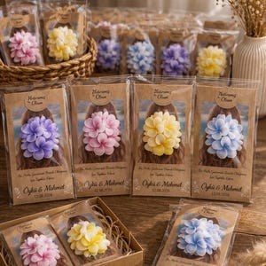 Può includere: Una collezione di accessori per capelli floreali in vari colori, tra cui rosa, giallo, blu e viola, confezionati in plastica trasparente e presentati in un cestino e una scatola. Ogni confezione ha un retro in carta marrone con testo e i nomi "Öykü & Mehmet" e la data 12.08.2026.