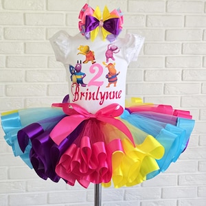 May include: A white short-sleeved shirt featuring cartoon characters, the number 2, and the name Brilynne in pink text. The shirt is paired with a multi-colored tulle tutu skirt with a pink ribbon bow and a matching hair bow in purple, yellow, and pink.