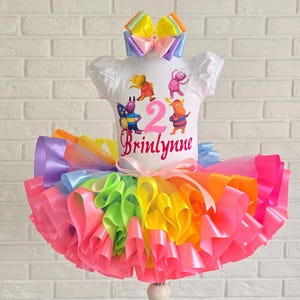 May include: A white short-sleeved shirt featuring a graphic of four cartoon characters, the number 2, and the name Brinlynne in pink script. The shirt is paired with a multi-colored rainbow tulle tutu skirt and a matching rainbow ribbon hair bow.