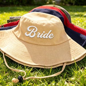 May include: A tan bucket hat with the word Bride embroidered in white script on the front. The hat features a wide brim, metal eyelets on the sides, and an adjustable chin strap with a black toggle. Additional hats in navy, red, and olive are stacked behind.