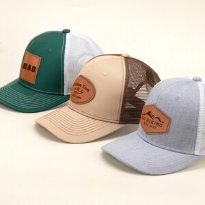 Könnte beinhalten: Drei Trucker-Caps mit Lederaufnähern. Eine ist grün mit "DAD" auf dem Aufnäher, eine andere ist beige mit "Custom Text" und einem Bergmotiv, und die dritte ist grau mit "GO HIKING" und einem Bergmotiv.