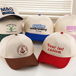 Puede incluir: Una colección de gorras de béisbol color crema con varios diseños y textos bordados. Las gorras presentan viseras de diferentes colores, incluyendo azul marino, verde, azul, rosa, marrón y rojo. Algunas gorras muestran texto como "M&G EST. 2025", "FROM AFAR", "custom embroidery", "mom Happy Birthday" y "Your text custom".