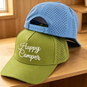 May include: Two baseball caps, one light blue and one green, stacked. The green cap features the embroidered words "Happy Camper" in white. Both caps have a perforated design. The caps are designed for outdoor activities.