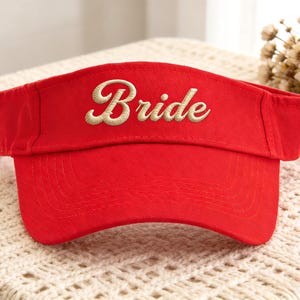 以下が含まれることがあります： 鮮やかな赤色のサンバイザー。前面には「Bride」の文字が金色の糸で刺繍されています。丈夫な生地で作られており、太陽から目を守るためのカーブしたつばが付いています。結婚式にぴったりのスタイリッシュなアクセサリーです。