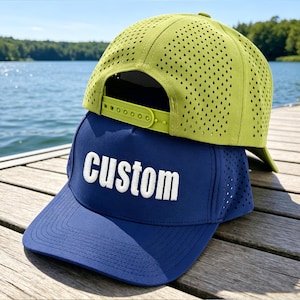 Könnte beinhalten: Eine limettengrüne, perforierte Snapback-Kappe, die auf einer marineblauen Baseballkappe liegt. Die marineblaue Kappe zeigt das Wort custom in weißen Blockbuchstaben auf der Vorderseite und hat Seitenteile mit kleinen Belüftungslöchern.