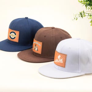 Könnte beinhalten: Drei Baseballkappen in Marineblau, Braun und Weiß. Jede Kappe hat ein quadratisches Lederpatch mit einem Design und Text. Die marineblaue Kappe hat ein Patch mit der Aufschrift "Since 1989". Die braune Kappe hat ein Patch mit einer Katze und einem Hund. Die weiße Kappe hat ein Bergdesign.