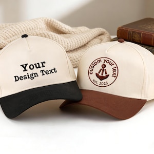 Op de afbeelding: Twee baseball caps met aangepaste tekstopties. Eén cap heeft een zwarte klep en de tekst "Your Design Text". De andere cap heeft een bruine klep met een ankerontwerp en de tekst "custom your text est. 2025". Beide caps zijn crèmekleurig.