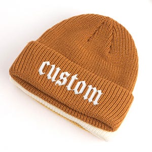 Könnte beinhalten: Eine senfgelbe Strickmütze mit umgeschlagenem Bündchen. Das Wort custom ist in weißem Garn in einer gotischen Schriftart auf die Vorderseite des Bündchens gestickt. Die Mütze hat eine durchgehende Rippenstruktur und ein weißes Innenfutter.