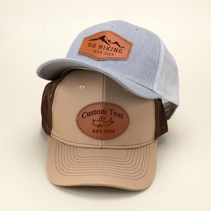 May include: Two baseball caps. The top cap is light blue with a white mesh back and a brown leather patch that says "GO HIKING EST. 2025". The bottom cap is tan with a brown mesh back and a brown leather patch that says "Custom Text EST. 2026".