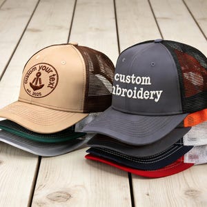 Puede incluir: Una pila de gorras de camionero en varios colores, incluyendo beige, gris y rojo. La gorra beige presenta un diseño de ancla bordada con el texto "custom your text est. 2025." La gorra gris tiene "custom embroidery" en blanco.
