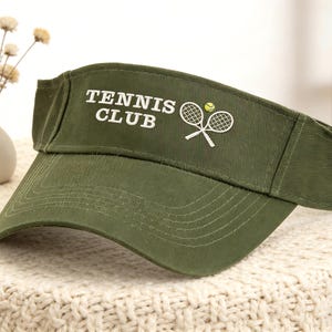 May include: An olive green tennis visor with the words "TENNIS CLUB" in white. It features a tennis ball and crossed rackets in white. The visor is made of fabric and is designed to shield the eyes from the sun. The visor is a sports accessory.