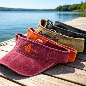 May include: A row of visors in various colors, including burgundy, orange, tan, and black, are displayed on a wooden dock. The burgundy visor has an embroidered sailboat design with the text "EST. 2026". The tan visor has the text "Custom Text".