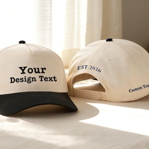 May include: Two cream-coloured baseball caps with black brims are shown. One cap has the text "Your Design Text" in black, while the other has "EST. 2026" and "Custom Text" in black. The caps are on a light-coloured surface.