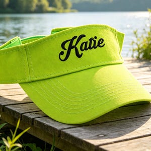 May include: A bright green visor with the name "Katie" embroidered in black script. The visor is made of a soft fabric and has a curved brim. It is sitting on a wooden surface with a lake in the background.