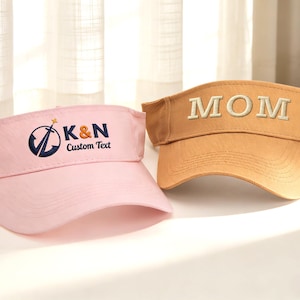 May include: Two visors are displayed. One is light pink with the text "K&N Custom Text" and a logo in navy blue. The other visor is tan with the embroidered word "MOM" in a lighter tan color. Both visors are made of a sturdy material.