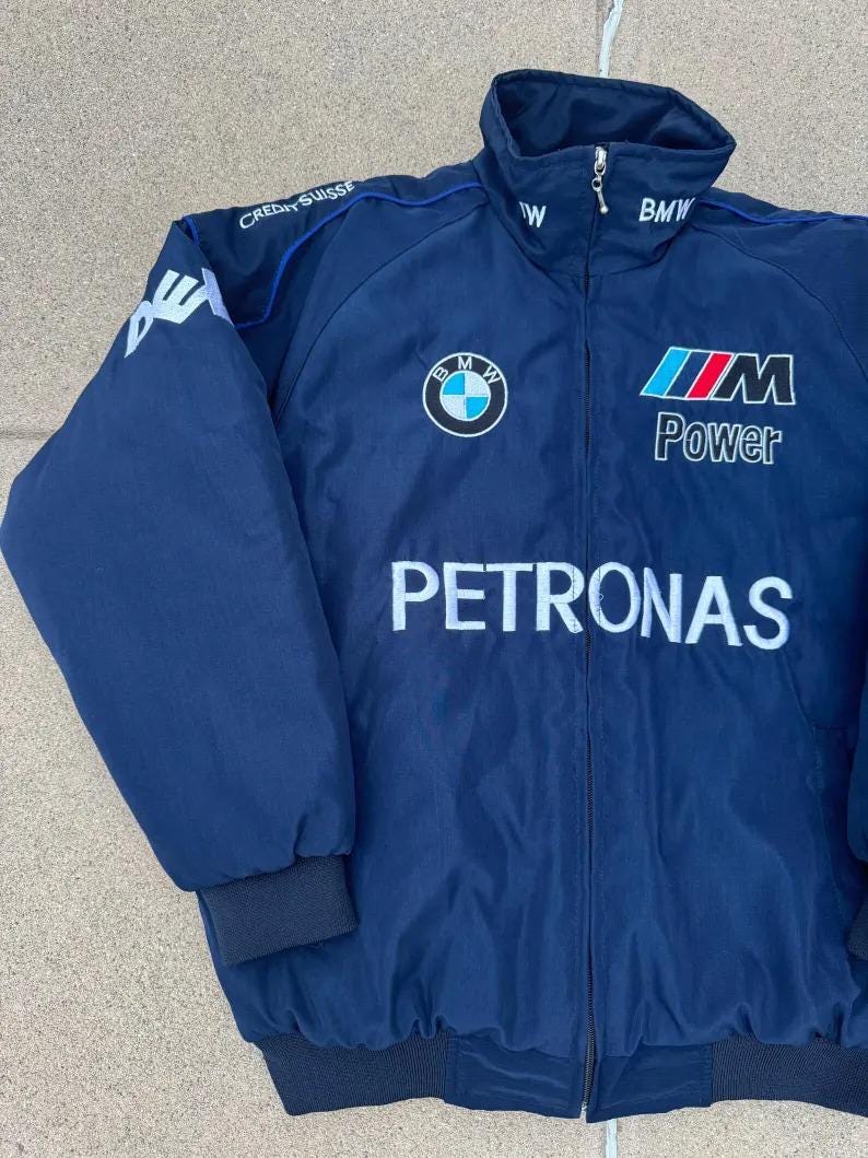 May include: Navy blue zip-up jacket with the BMW logo, "M Power", and "Petronas" text. The jacket has a stand-up collar, blue trim, and black cuffs and hem. The left sleeve has a white logo and the text "Credit Suisse".
