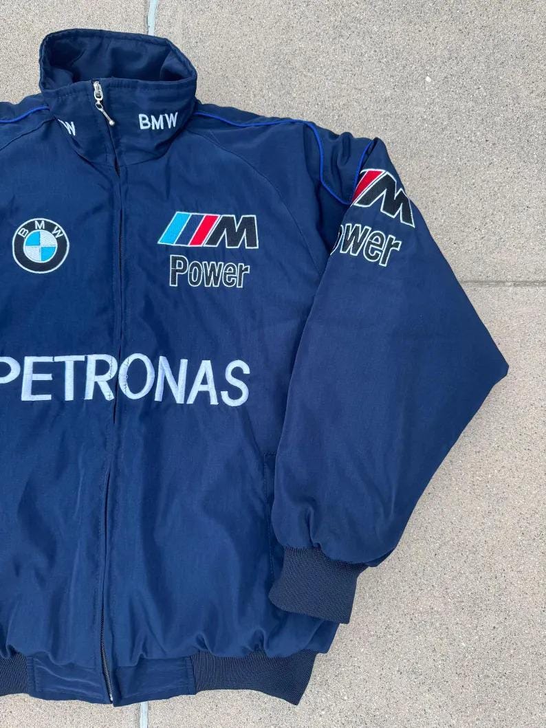 May include: Navy blue BMW Petronas jacket with a high collar and a zipper closure. The jacket features the BMW logo, the words "BMW", "Power", and "Petronas" in white, red, and blue. The sleeves have black cuffs.
