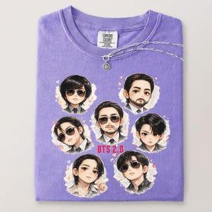 May include: A lavender Comfort Colors t-shirt featuring cartoon portraits of seven people wearing sunglasses. The text "BTS 2.0" is below the portraits. A silver necklace with a pendant is draped across the shirt.