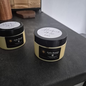Puede incluir: Dos frascos de pomada para cabello y cuero cabelludo Kaks Beauty con tapas y etiquetas negras. Las etiquetas dicen "Handmade with love" y "Kakra". Los frascos están sobre una superficie gris oscuro.