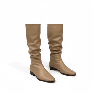 May include: A pair of beige knee-high boots with a slightly wrinkled texture. The boots have a pointed toe and a low, black heel. The boots are made of a smooth material and are set against a white background.