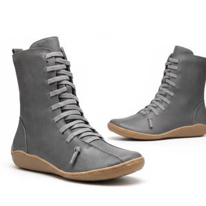 K&ouml;nnte beinhalten: Zwei graue Lederstiefel mit hellbraunen Sohlen. Die Stiefel haben eine mittlere Wadenh&ouml;he und sind vorne mit grauen Schn&uuml;rsenkeln geschn&uuml;rt. Das Design ist minimalistisch und modern, passend f&uuml;r verschiedene Outfits.