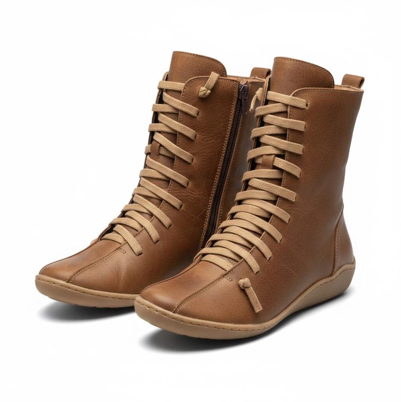 K&ouml;nnte beinhalten: Ein Paar braune Lederstiefel mit beigen Schn&uuml;rsenkeln und einem seitlichen Rei&szlig;verschluss. Die Stiefel haben eine runde Spitze und eine flache Sohle. Die Stiefel sind kniehoch.