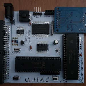 Può includere: Una scheda di circuito grigio chiaro con vari componenti elettronici, tra cui chip, connettori e un modulo blu. Il testo sulla scheda include "UL.I.f.A.C." e "Ultimate Interface for Amstrad CPC."
