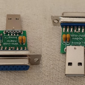 May include: Two 15-pin to USB adapters featuring green circuit boards with silver metal connectors, blue plastic ports, and small yellow capacitors. The boards are printed with the text 15Pin->USB Adapter ikonsgr74 in white lettering.