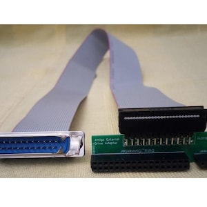 Könnte beinhalten: Ein Amiga-Externlaufwerkadapter mit einem grauen Flachbandkabel, einem blauen und silbernen Stecker und einer grünen Leiterplatte. Die Leiterplatte trägt den Text "Amiga External Drive Adapter" und "Data Connector".