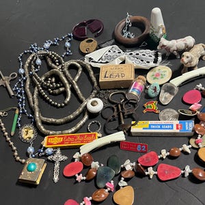 Puede incluir: Una colección de joyas vintage y pequeños objetos. Los artículos incluyen collares, cuentas, un colgante de cruz, un libro pequeño, una caja de "LEAD" y otros objetos decorativos. La paleta de colores incluye plata, marrón, rojo y azul.