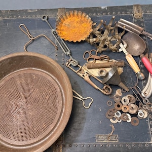 Puede incluir: Un surtido de herramientas y utensilios de cocina antiguos de metal, que incluyen un colador grande, un molde para tartas y varias pinzas y cucharas. Los artículos están dispuestos sobre una superficie oscura y texturizada.