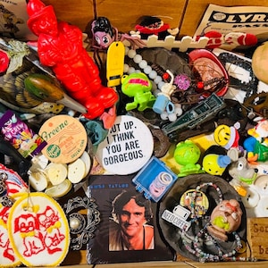 May include: A collection of assorted items including a bright red plastic firefighter figure, a white button with the text SHUT UP, I THINK YOU ARE GORGEOUS, a James Taylor album cover, a green frog toy, various beads, patches, and small trinkets.